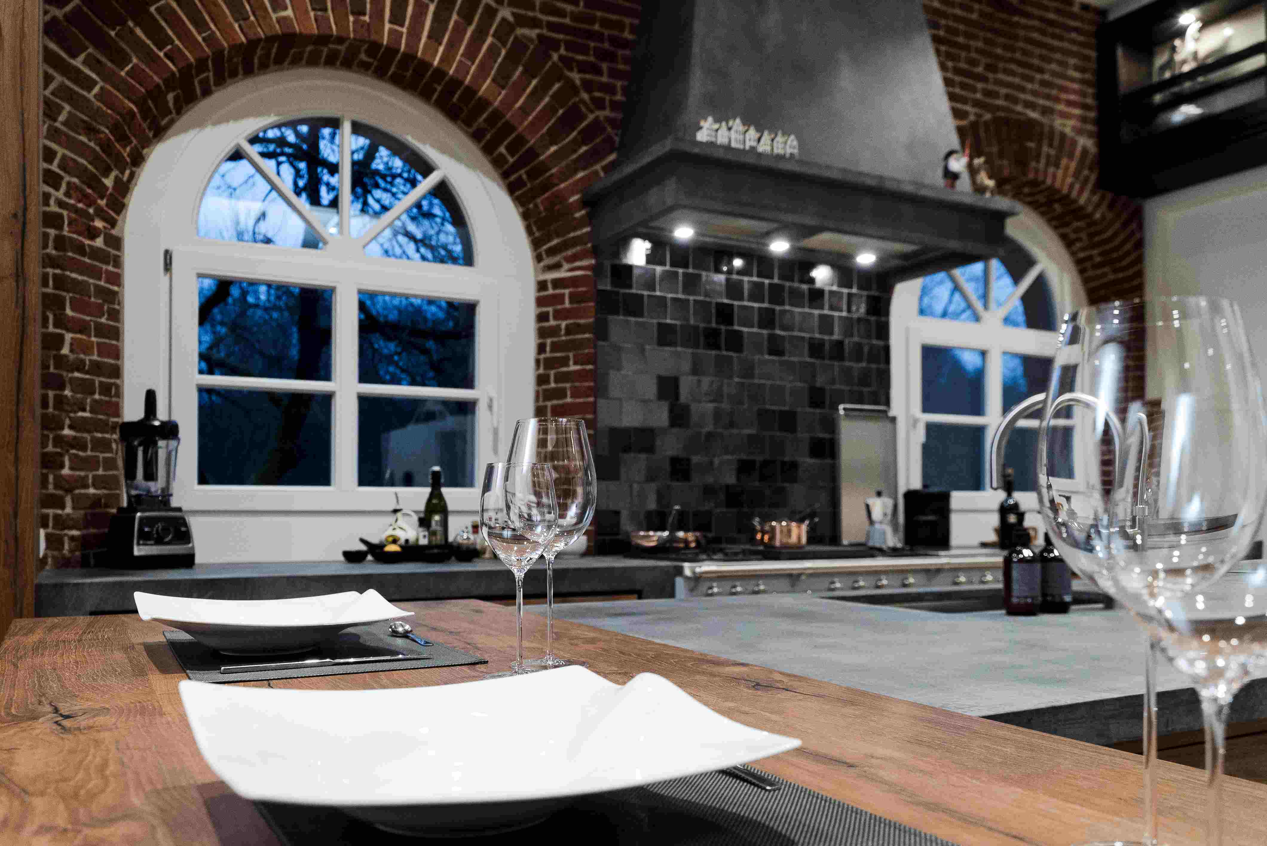 Innenansicht einer modernen Küche mit einem rustikalen Holztisch, gedeckt mit eleganten weißen Tellern und Weingläsern, vor einem großen Fenster mit Ziegelbogen.