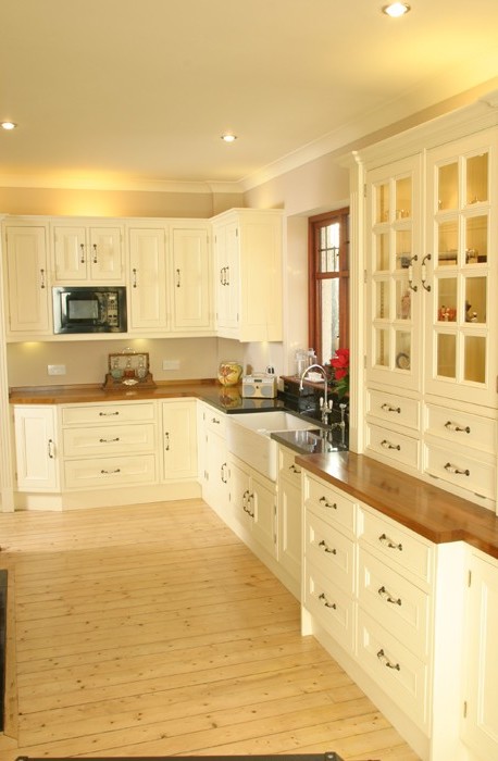 Moderne Landhausküche mit cremefarbenen Holzmöbeln und Echtholz-Arbeitsplatte Gemütliche Landhausküche mit cremefarbenen Schränken, Holzarbeitsplatte, Einbaumikrowelle und Spüle vor einem Fenster