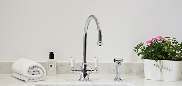 Eleganter Wasserhahn mit Doppelgriff und moderner Armatur auf weißer Marmor-Arbeitsplatte Moderner Wasserhahn aus Chrom mit zwei Griffen, daneben eine weiße Schale mit rosa Blumen, ein Handtuch und Seifenspender auf einer Marmorfläche