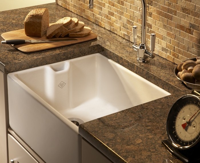 Moderne Landhausküche mit weißem Keramikspülbecken, brauner Arbeitsplatte und Brotschneidebrett Küchenspüle aus weißer Keramik mit brauner Stein-Arbeitsplatte, daneben ein Brotschneidebrett mit aufgeschnittenem Brot und Messer