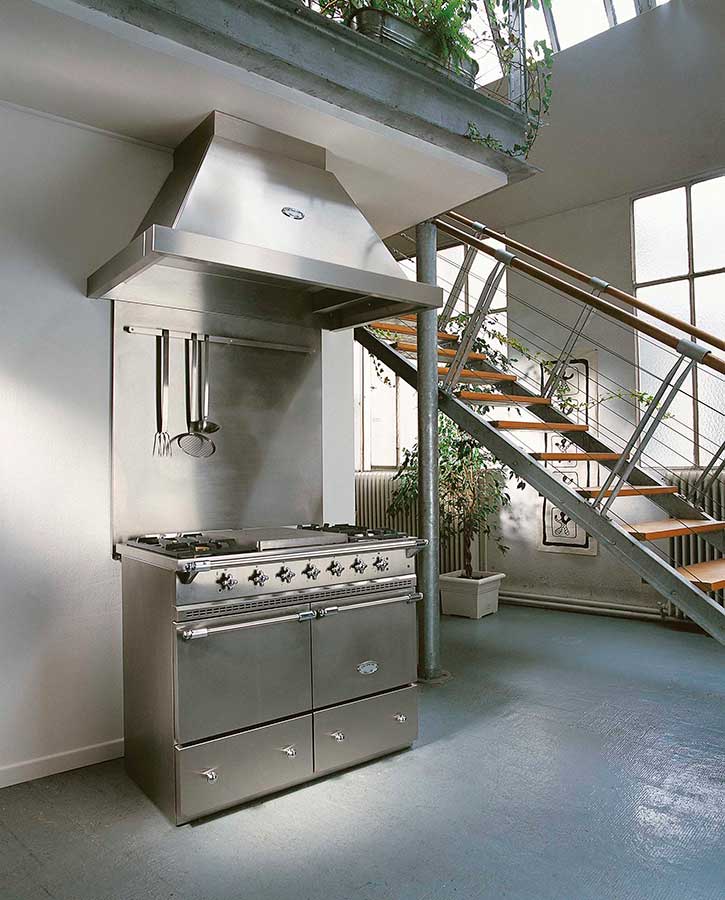 Professioneller Edelstahl-Gasherd mit Dunstabzugshaube in moderner Loftküche Großer Edelstahl-Gasherd mit passender Dunstabzugshaube und Küchenutensilien in einer offenen, lichtdurchfluteten Loftküche neben einer Treppe