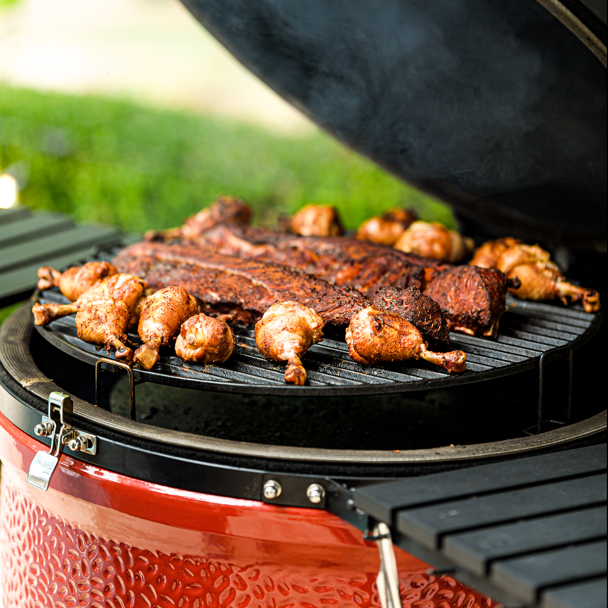 Saftige Hähnchenschenkel und Rippchen auf einem Keramikgrill im Garten – BBQ Genuss für Grillfreunde Geöffneter Keramikgrill mit knusprigen Hähnchenschenkeln und herzhaften Rippchen auf dem Grillrost, im Hintergrund grüner Garten