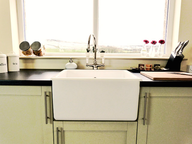 Moderne Landhaus-Küche mit Keramik-Spülbecken und stilvoller Arbeitsplatte Helle Küche mit großem weißen Keramik-Spülbecken, Armatur, schwarzen Arbeitsflächen, Vorratsgläsern, Messerblock und Blick aus dem Fenster auf die Landschaft