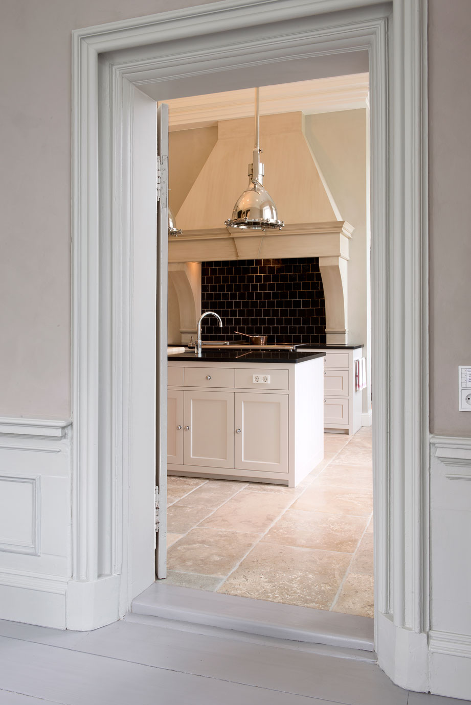Elegante Landhausküche mit Kücheninsel, schwarzen Fliesen und modernen Hängelampen Blick durch eine offene Tür auf eine stilvolle Landhausküche mit Kücheninsel, dunkler Fliesenrückwand, Unterbauwaschbecken und großen silbernen Hängelampen