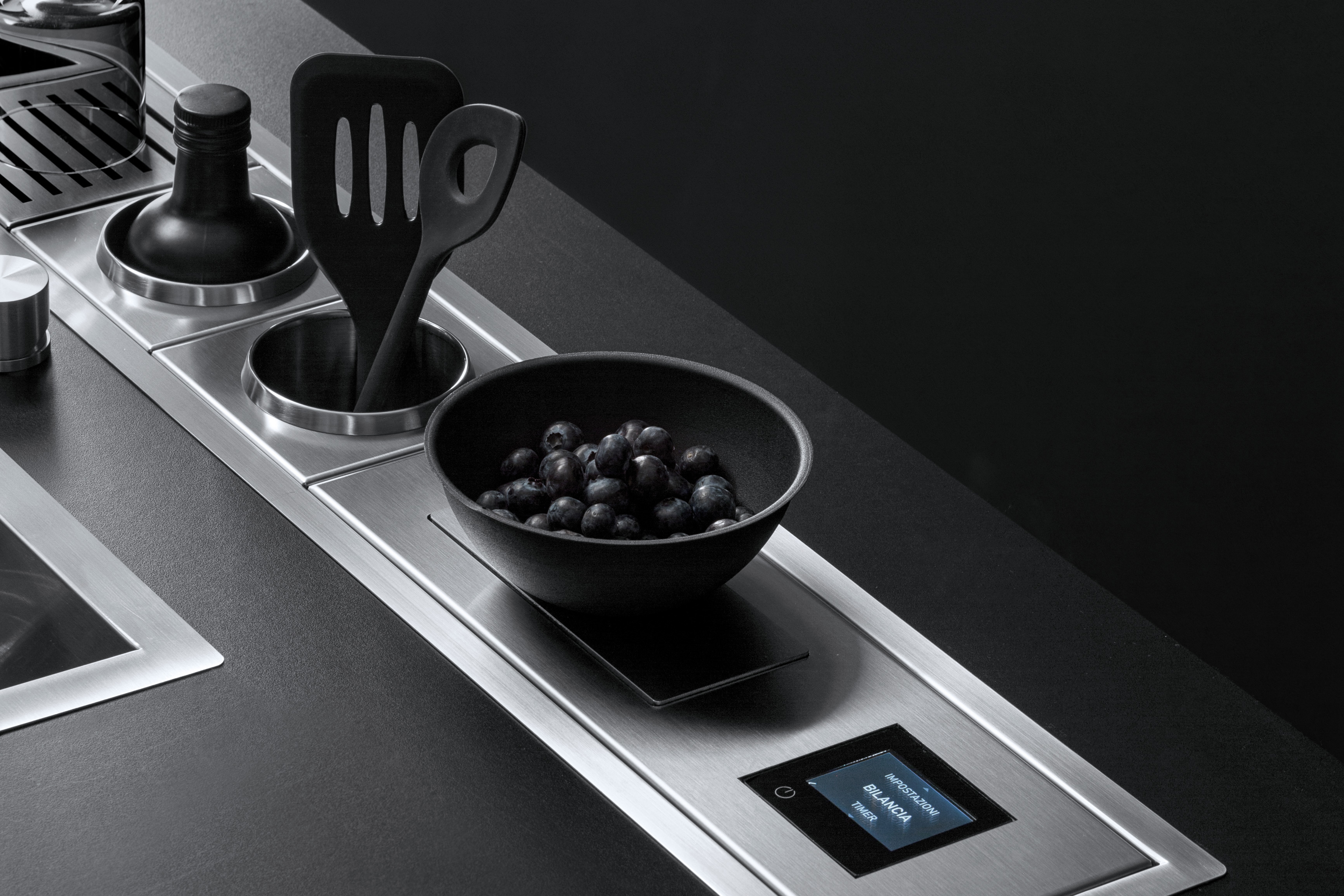Moderne Küchenzeile mit integrierten Edelstahl-Elementen, Schale mit Heidelbeeren und Touchdisplay Detailansicht einer modernen Küchenarbeitsplatte mit Edelstahl-Elementen, schwarzer Schale voller Heidelbeeren und Digitaldisplay