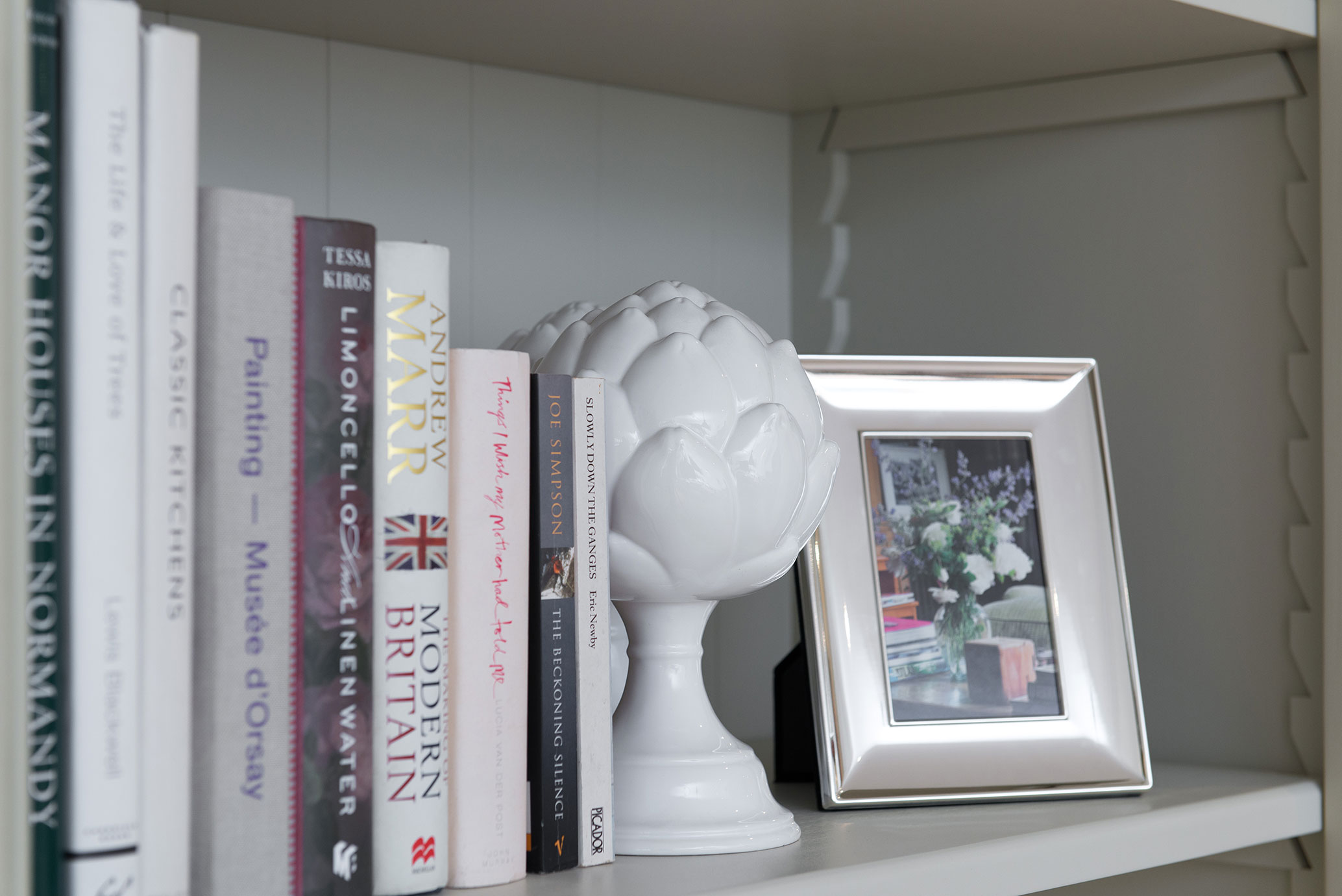 Bücherregal mit dekorativer Keramik-Artischocke und Fotorahmen Nahaufnahme eines Bücherregals mit verschiedenen Büchern, einer weißen Keramik-Artischocke und einem silbernen Fotorahmen mit Blumenbild.
