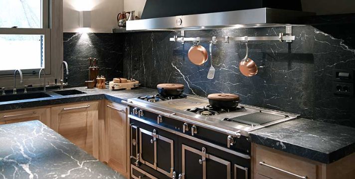 Hochwertiger Range Cooker/Gasherd in L-Form-Küche mit schwarzer Naturstein-Arbeitsplatte, Edelstahlhaube, Hängeschiene und Kupfertöpfen – ideal für Premium-Küchenausstattung