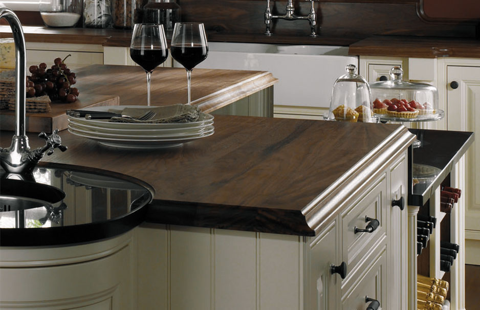 Hochwertige Küchenarbeitsplatte aus Holz in klassischer Landhausküche Edle Küchenarbeitsplatte aus dunklem Massivholz, gedeckter Tisch mit Tellern und Weingläsern, im Hintergrund Obst und Gebäck in einer stilvollen Landhausküche.