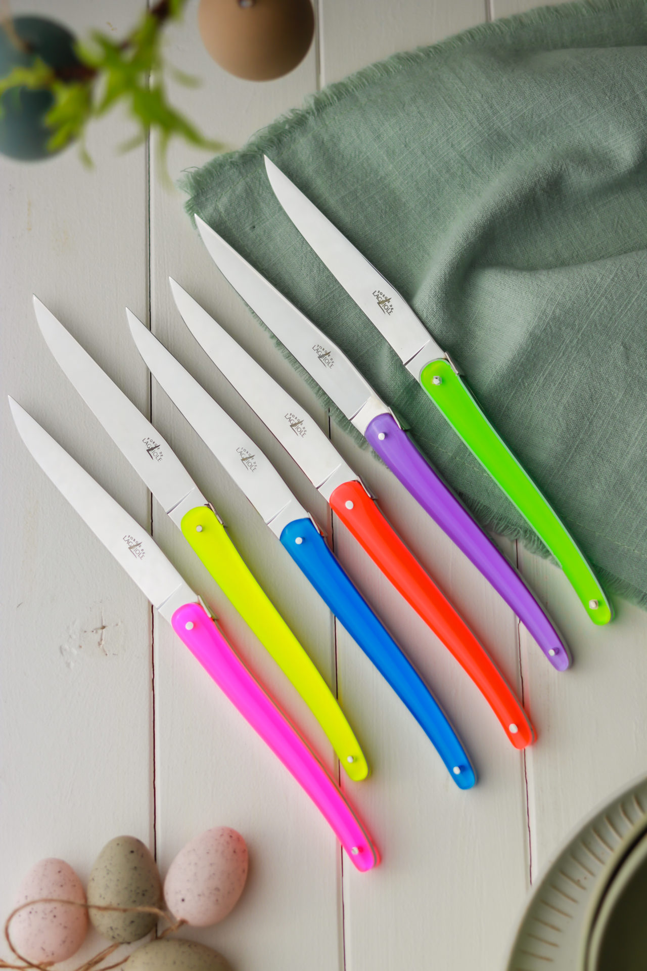 Set aus sechs bunten Steakmessern mit Edelstahlklingen – farbenfrohe Messer für den Esstisch Sechs Steakmesser mit bunten Griffen in Neonfarben liegen nebeneinander auf einem hellen Holztisch neben einer grünen Serviette und Ostereiern.