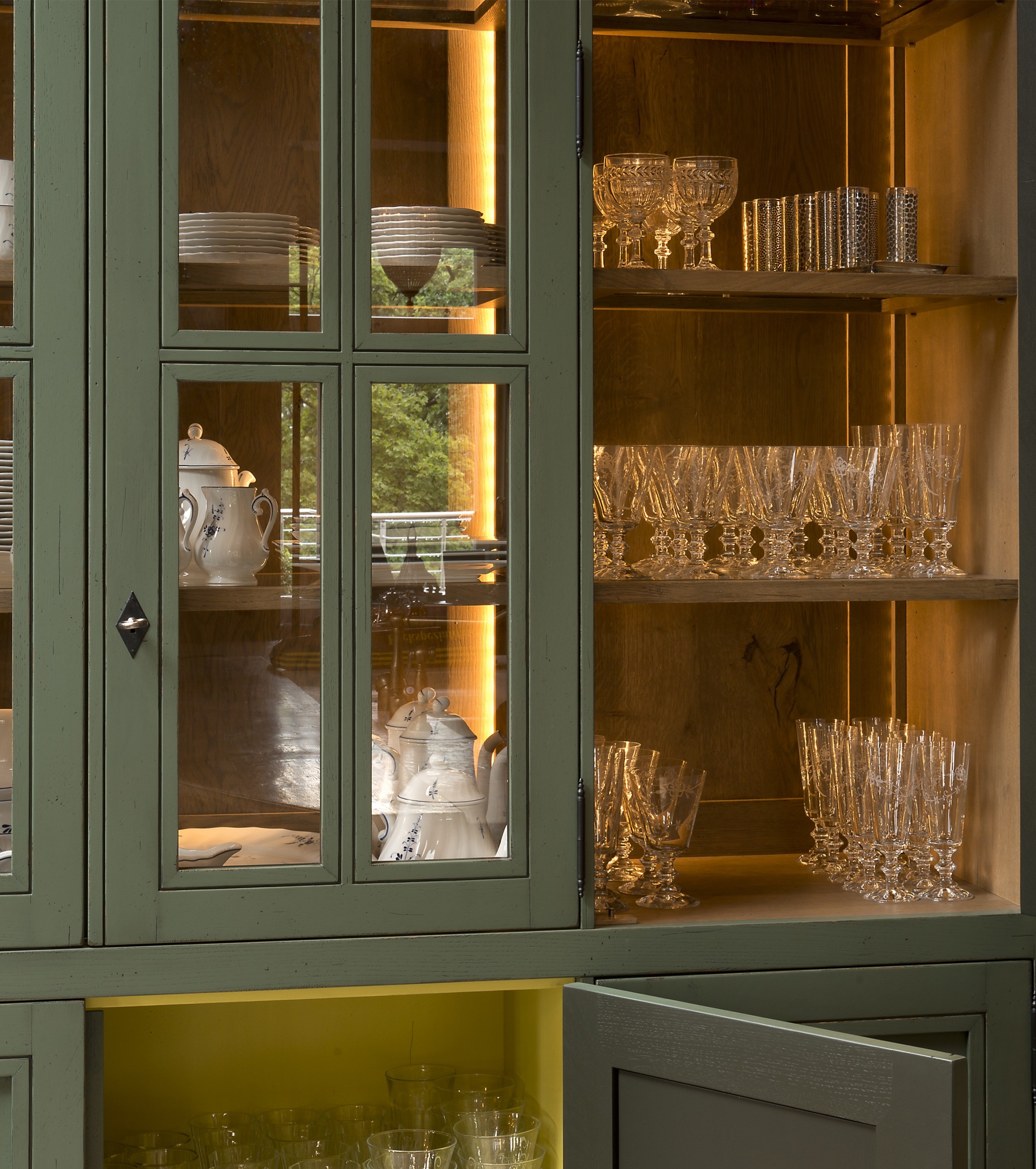 Elegante Vitrine mit Porzellangeschirr und Kristallgläsern, beleuchtet in einem grünen Küchenschrank