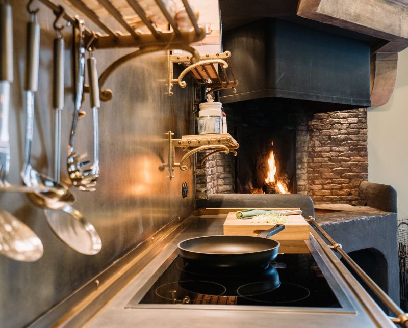 Moderne Küche mit Induktionskochfeld, Pfanne und offenem Kamin für gemütliches Kocherlebnis Hochwertige Küche mit Hängeregalen, Kochutensilien, einem Induktionskochfeld mit Pfanne und einem traditionellen Kamin im Hintergrund