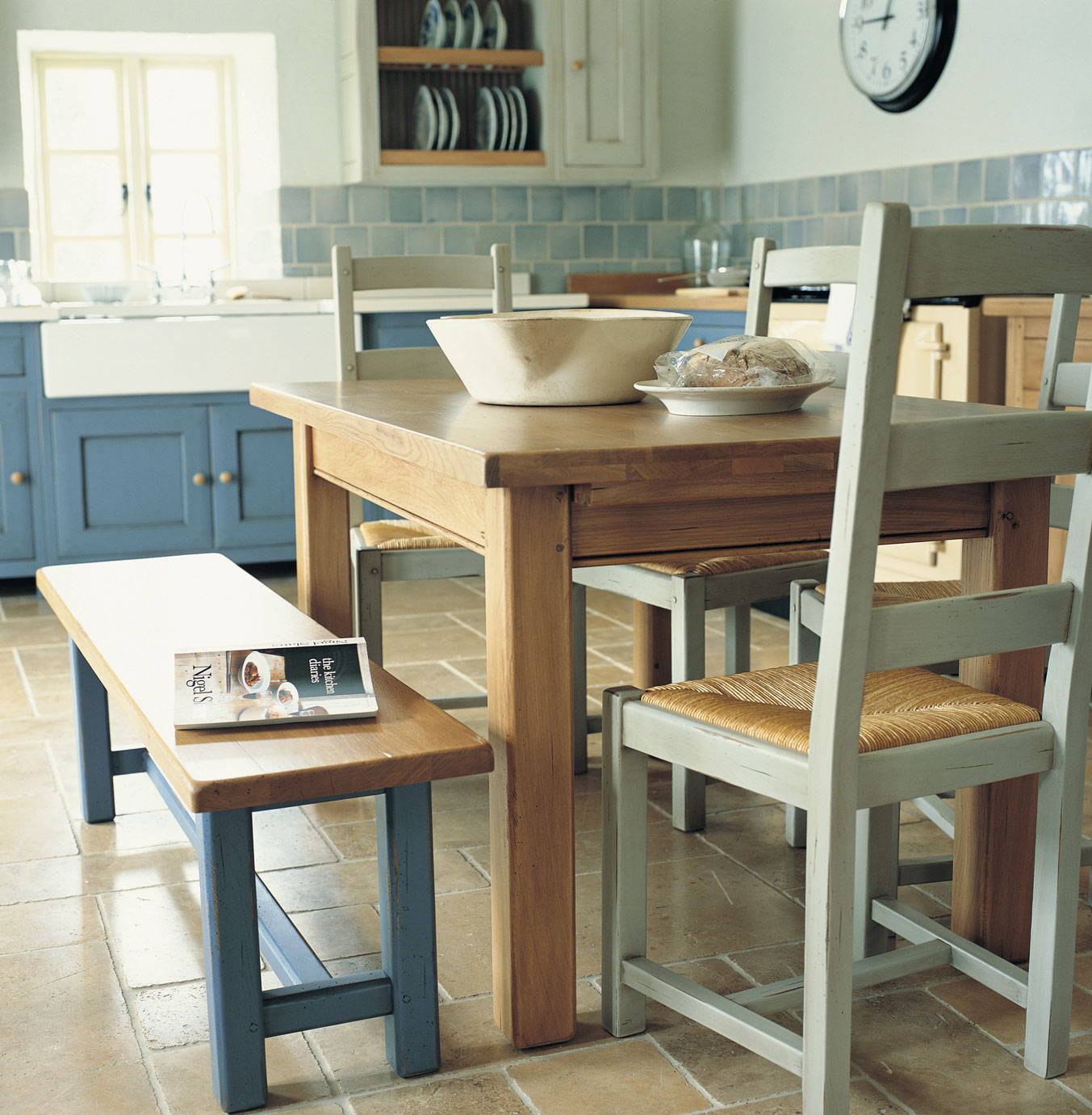 Esszimmer im Landhausstil mit Holztisch, Holzbank und Stühlen auf Fliesenboden in heller, einladender Küche Holztisch mit Holzbank und Stühlen in einer freundlichen Landhausküche mit blauen Schränken und Keramikschale auf dem Tisch