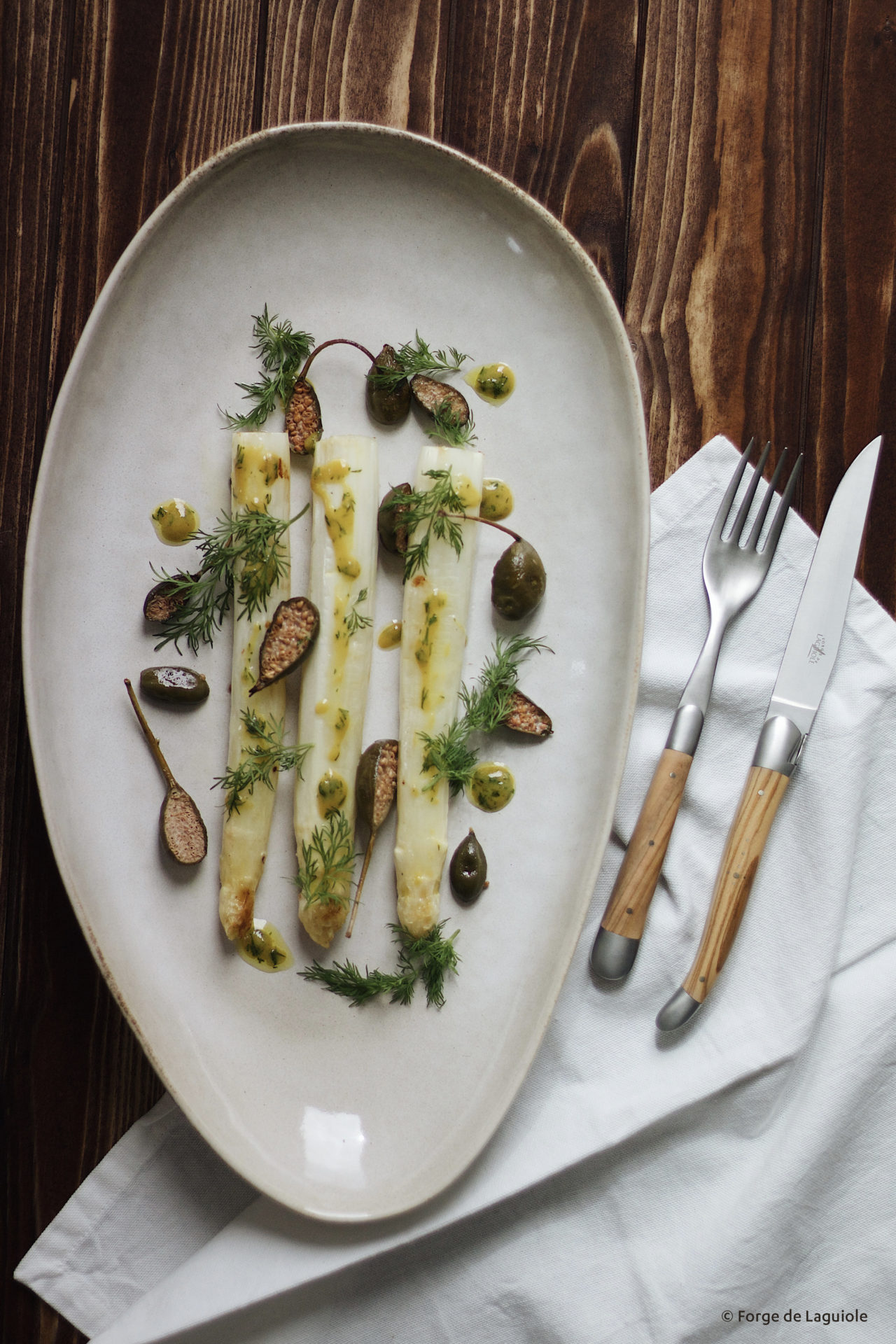 Gourmet-Teller mit weißem Spargel, Kapern, frischen Kräutern und Dressing auf hochwertigem Porzellan Servierteller mit drei Stangen weißem Spargel, Kapern, frischem Dill und Kräuterdressing, daneben edles Besteck auf einer Serviette