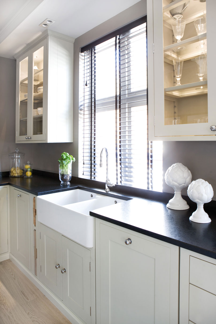Moderne, helle Küche mit Spülbecken, Glastürenschränken und schwarzen Arbeitsflächen Helle Küchenzeile mit großem weißen Spülbecken, schwarzen Arbeitsplatten, dekorativen Gläsern und Fenstern mit Jalousien