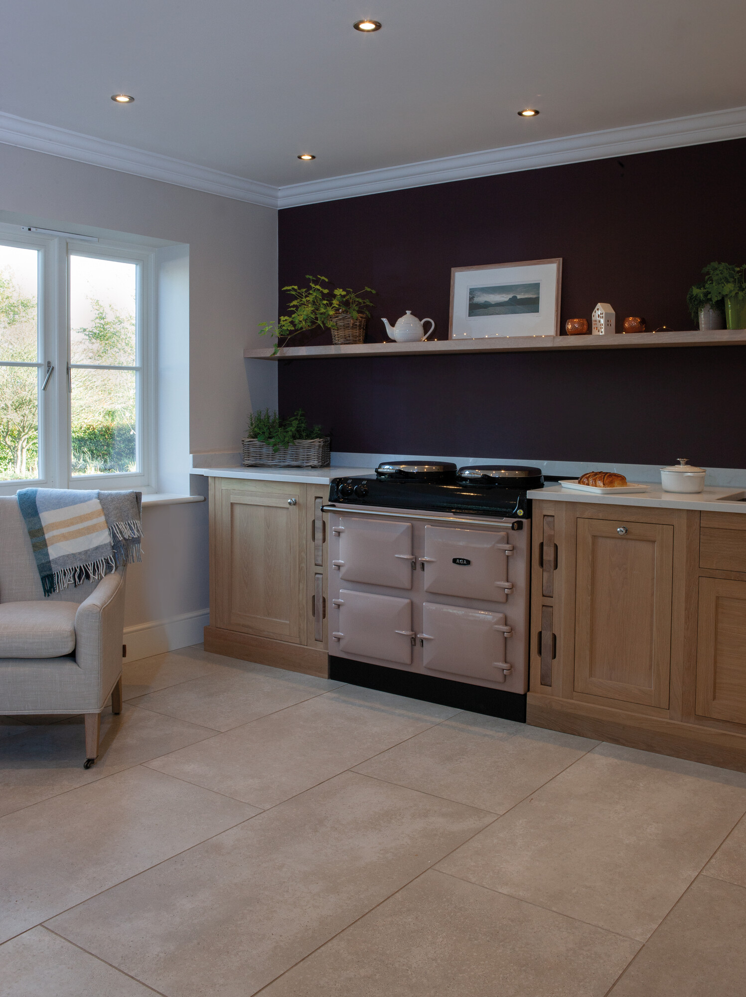 Moderne Landhausküche mit cremefarbenem AGA-Herd, Holzmöbeln und dekorativer Küchenzeile Gemütliche Küche mit cremefarbenem AGA-Herd, hellen Holzmöbeln, dunkler Wand, Fenster, Pflanzen und schlichtem Dekor